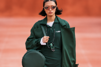 Silhouette verte du défilé Lacoste printemps-été 2027 sur podium terracotta, avec manteau long, mini-robe, lunettes de soleil et sac rond.