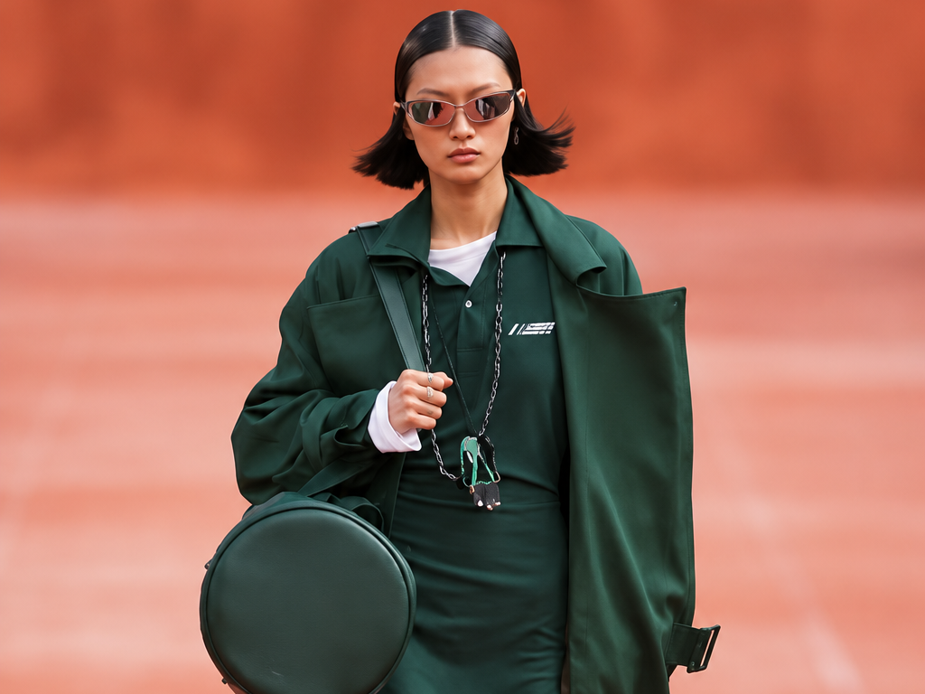 Silhouette verte du défilé Lacoste printemps-été 2027 sur podium terracotta, avec manteau long, mini-robe, lunettes de soleil et sac rond.