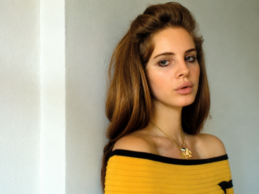 Lana Del Rey dans une scène inspirée du clip Video Games, portant un haut jaune aux bordures noires et un pendentif doré devant un mur clair.