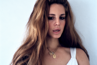 Lana Del Rey dans le clip Video Games, cheveux longs volumineux, maquillage rétro et robe blanche, dans une esthétique vintage.