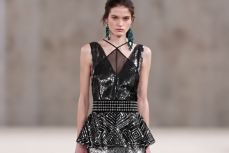 Mannequin du défilé Lanvin printemps-été 2027 portant une longue robe noire à sequins argentés, taille structurée, basque géométrique et boucles d'oreilles vertes.