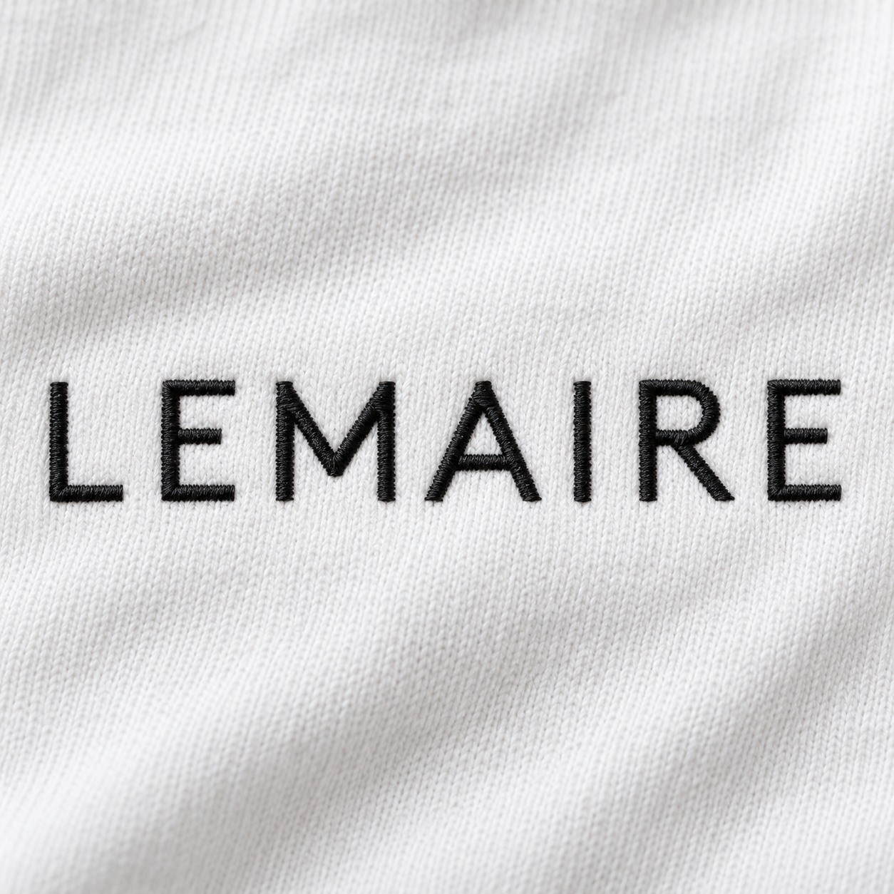 Logo Lemaire brodé en fil noir sur un tissu blanc en coton, détail textile de la maison de mode française.