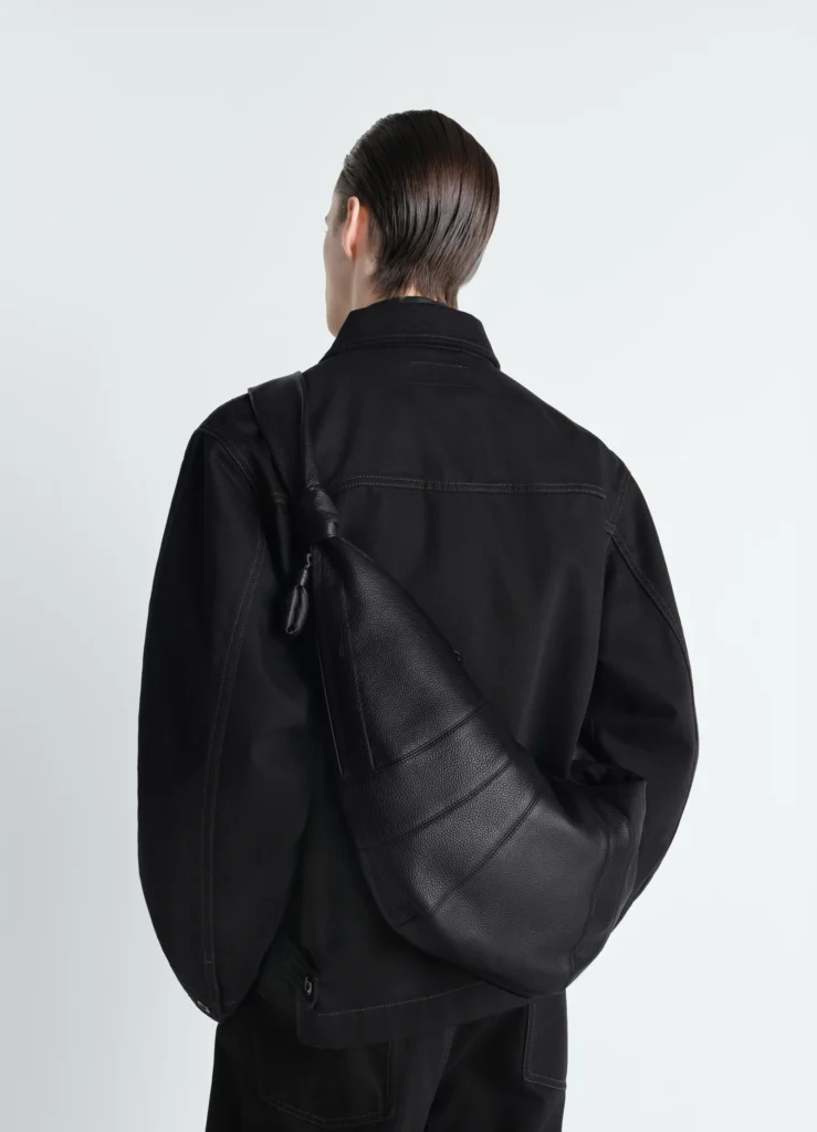 Sac Croissant Lemaire en cuir noir grainé, porté à l'épaule sur une veste noire, photographié de dos.