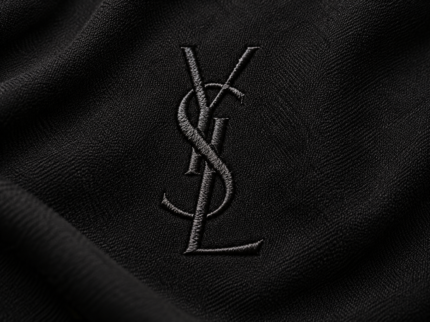 Logo YSL Yves Saint Laurent brodé en noir sur un tissu noir, détail du monogramme historique aux lettres entrelacées.