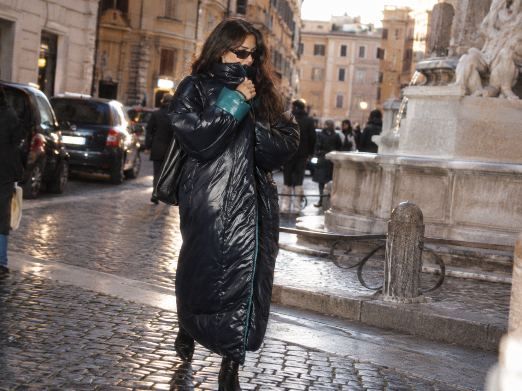 Jeune femme brune portant un Sleeping Bag Coat Norma Kamali noir brillant, long jusqu'aux chevilles, dans une rue pavée de Rome.