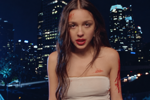 Olivia Rodrigo dans le clip Vampire, vêtue d'une robe blanche tachée de sang devant les immeubles illuminés de Los Angeles.