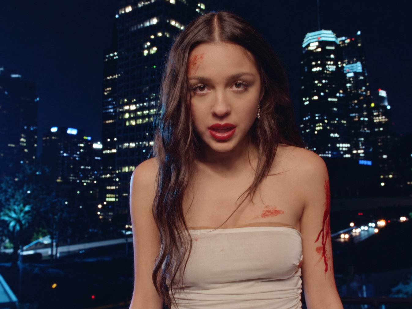 Olivia Rodrigo dans le clip Vampire, vêtue d'une robe blanche tachée de sang devant les immeubles illuminés de Los Angeles.
