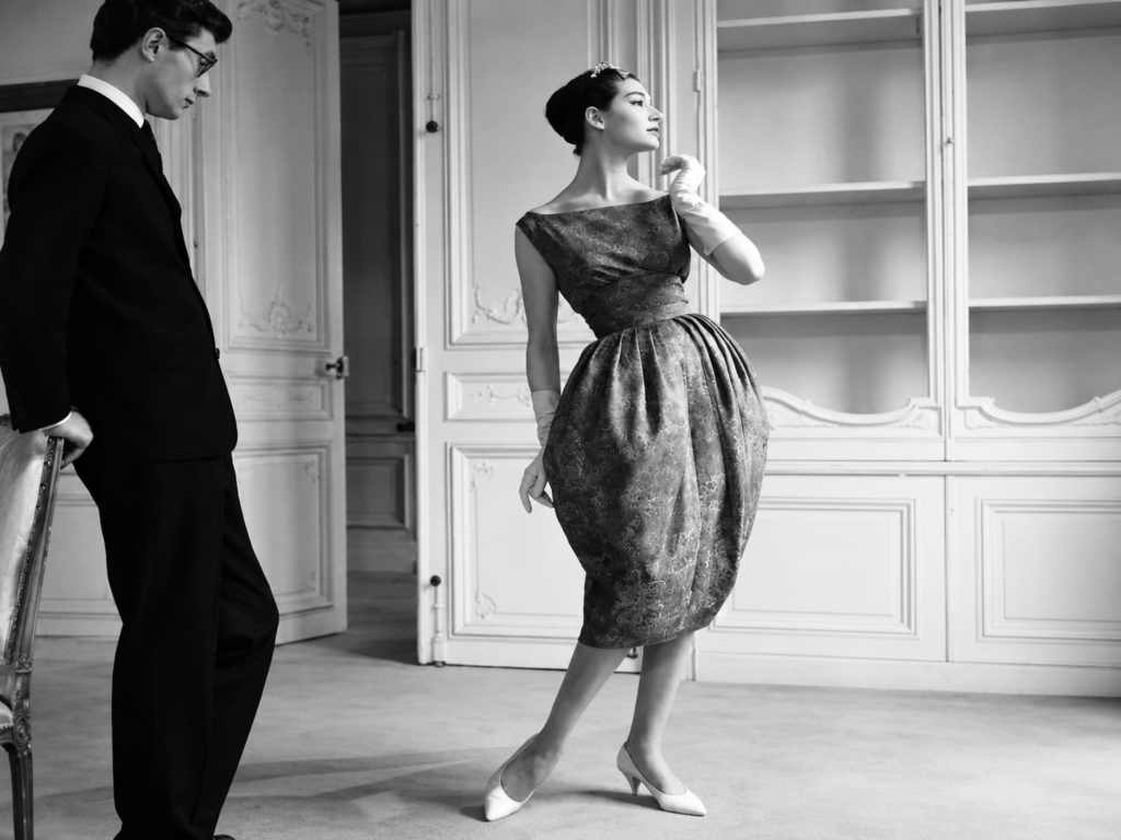 Scène de haute couture en noir et blanc montrant un mannequin en robe sculpturale, observé par un homme en costume.