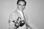 Pierre Cardin en costume clair, tenant des objets métalliques perforés, dans un portrait en noir et blanc.