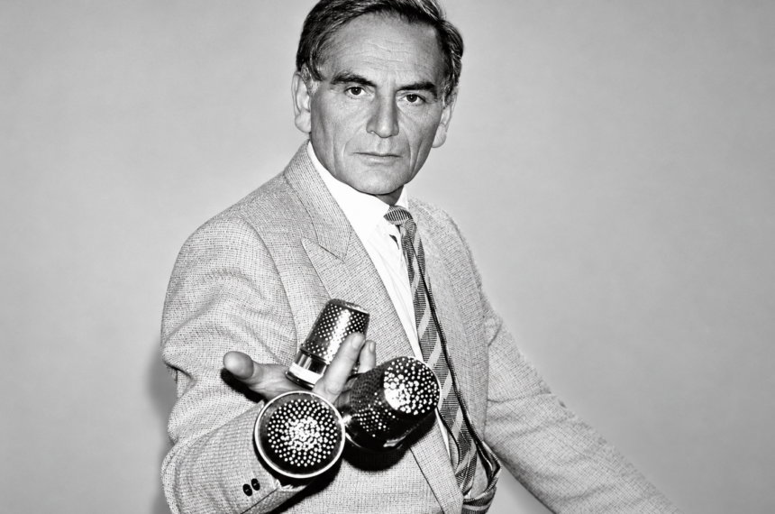 Pierre Cardin en costume clair, tenant des objets métalliques perforés, dans un portrait en noir et blanc.
