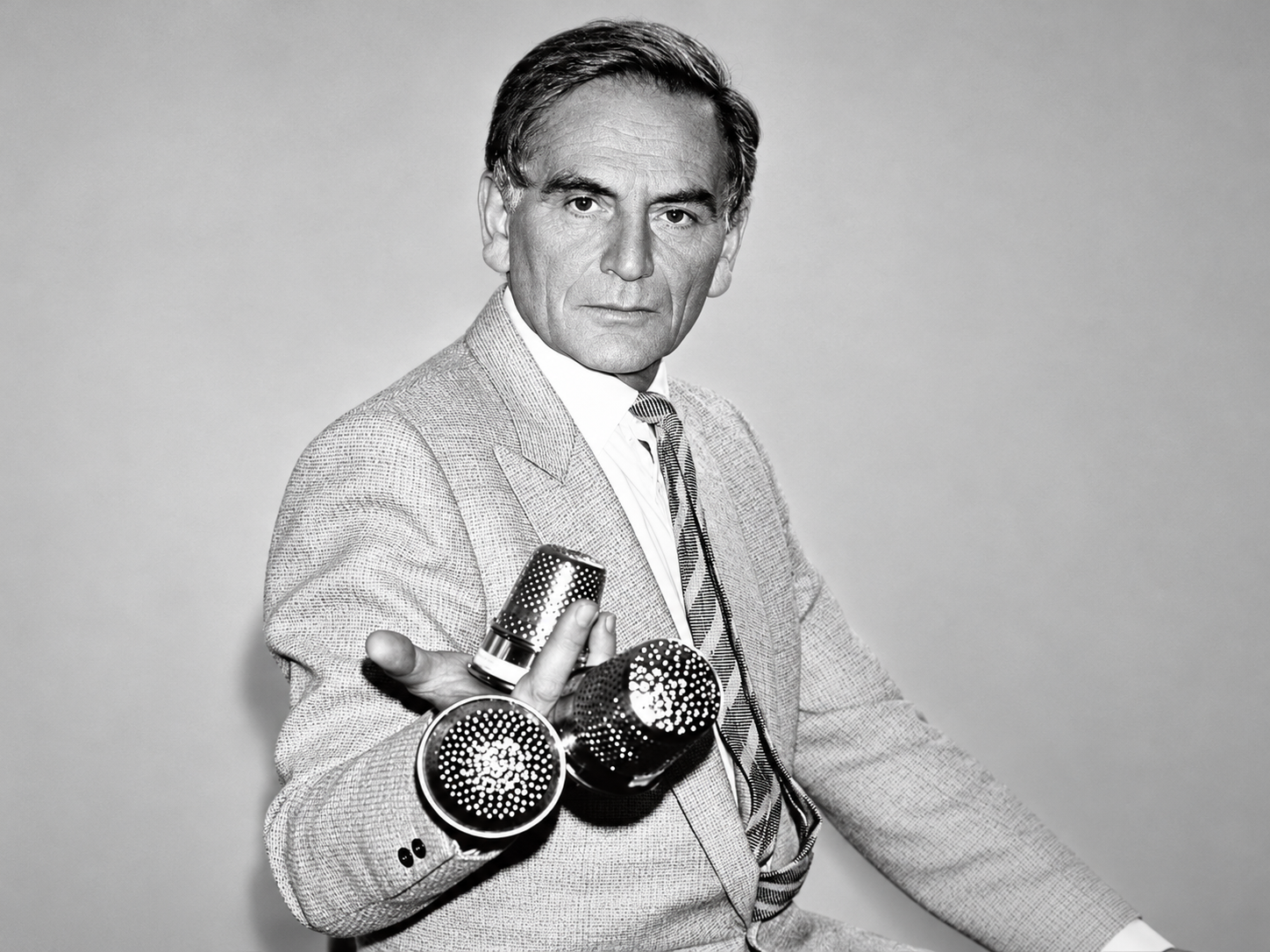 Pierre Cardin en costume clair, tenant des objets métalliques perforés, dans un portrait en noir et blanc.