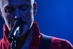 Josh Homme, chanteur de Queens of the Stone Age, au micro sous un éclairage bleu, vêtu d'une veste rouge.