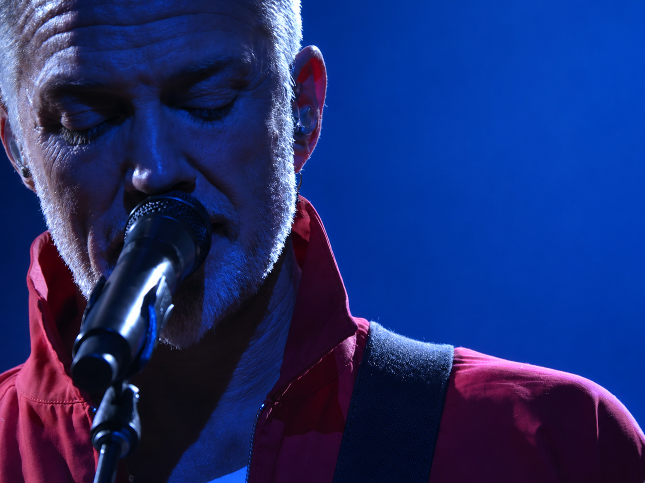 Josh Homme, chanteur de Queens of the Stone Age, au micro sous un éclairage bleu, vêtu d'une veste rouge.