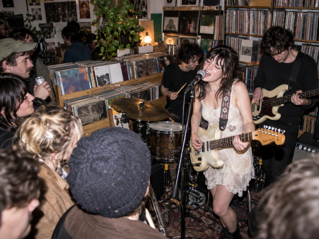 Une chanteuse joue de la guitare avec son groupe devant un public réuni pour un showcase dans un magasin de vinyles.