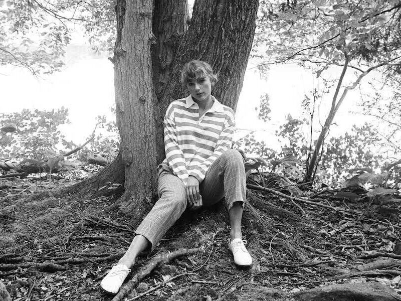Taylor Swift assise contre un arbre en chemise rayée et pantalon à carreaux pour l’univers de l’album Folklore.
