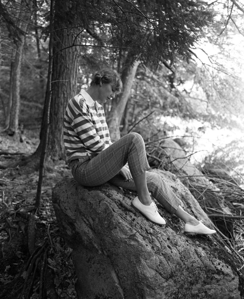 Taylor Swift assise sur un rocher en forêt, en chemise rayée et pantalon à carreaux dans l’univers de Folklore.