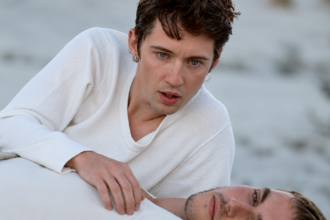 Troye Sivan, cheveux bruns et pull blanc, penché au-dessus d’un homme allongé dans une scène aux tons froids.