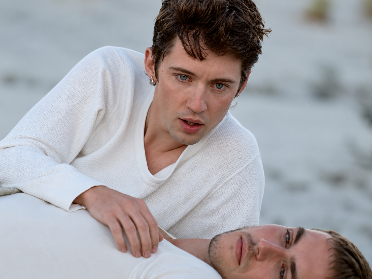 Troye Sivan, cheveux bruns et pull blanc, penché au-dessus d’un homme allongé dans une scène aux tons froids.