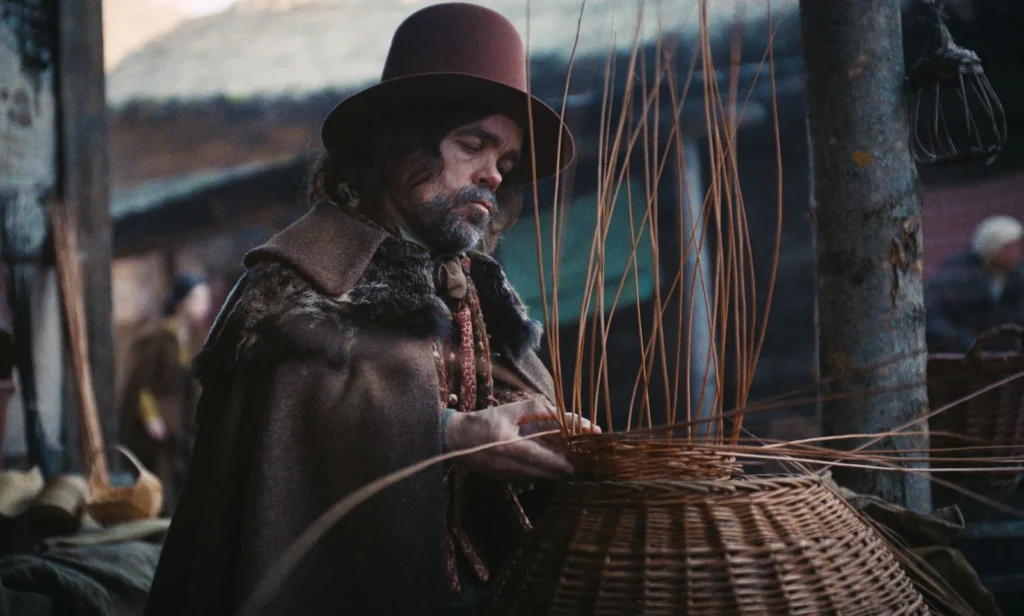 Peter Dinklage dans Wicker, coiffé d’un chapeau brun et vêtu d’un manteau épais, travaillant un panier en osier.